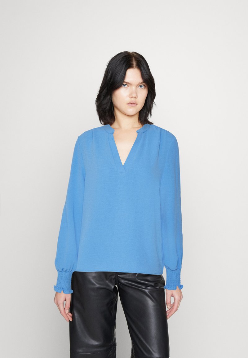 Top azzurro chiaro a maniche lunghe con scollatura a V e polsini arricciati, abbinato a pantaloni di pelle neri. Tessuto morbido e vestibilità comoda.