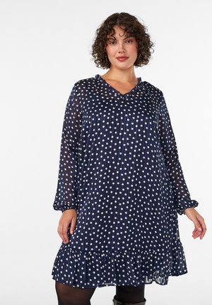 Donna che indossa un vestito blu navy a maniche lunghe con pois bianchi e orlo con balze, in piedi davanti a uno sfondo bianco semplice.