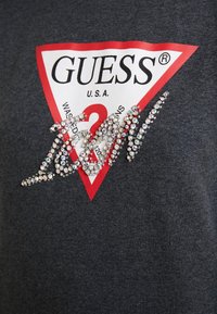 Camiseta gris de algodón con un gran logotipo triangular rojo y blanco que incluye el texto "GUESS", adornado con piedras de strass transparentes en un diseño cursivo.