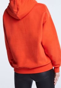 Sudadera con capucha naranja de corte holgado, costuras visibles y puños acanalados. La combinación se lleva con pantalones negros ajustados, que muestran una textura suave.