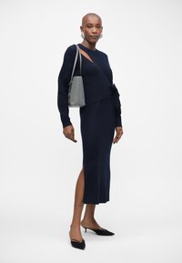 Vestido de malha azul marinho com mangas longas, apresentando um corte lateral e um detalhe de laço na cintura; combinado com uma bolsa cinza e sapatos de salto pretos.