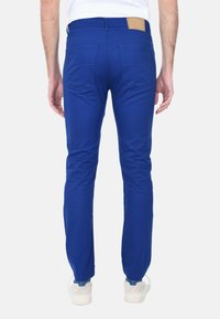 Jeans in denim blu con una vestibilità slim, due tasche posteriori e una patch in pelle beige sulla vita. Il tessuto sembra liscio e leggermente elasticizzato.