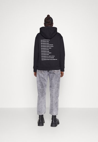 Black hoodie with white text on the back, paired with light gray denim jeans and black ankle boots. Cotton material, relaxed fit, and hooded design.
