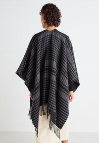 Schwarzer und grauer karierter Poncho mit Fransen, aus einem weichen Material. Verfügt über ein lockeres, fließendes Design und weit geschnittene Ärmel.