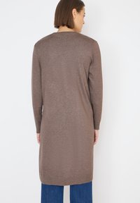 Bruine lange mouwen gebreide jurk met een ronde hals, rechte snit en een iets langere achterkant, gecombineerd met blauwe jeans.