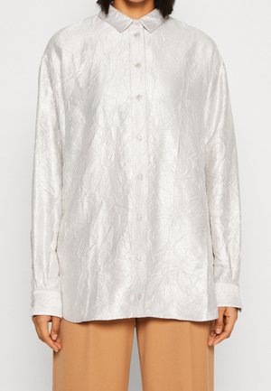 Vrouw draagt een wit, gekreukeld, blouse met lange mouwen en puntige kraag en beige wijde broek, staand tegen een effen achtergrond.
