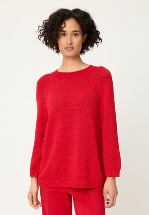 Femme aux cheveux bouclés et foncés portant un pull en maille rouge ample et un pantalon rouge assorti, debout devant un fond clair uni.
