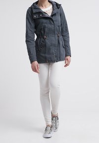 Chaqueta gris oscuro con capucha, cintura ajustada y bolsillos. Llevada sobre jeans pitillo blancos y zapatos tipo slip-on con estampado de serpiente.