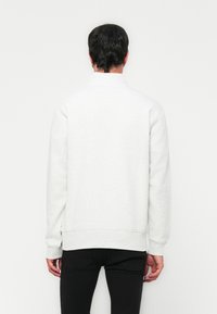 Homme portant un sweat-shirt blanc uni à manches longues et un pantalon noir, se tenant face à un mur blanc avec les mains détendues sur les côtés.