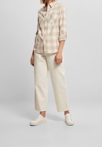 Femme portant une chemise à carreaux beige et blanche boutonnée, un pantalon large blanc cassé, et des baskets blanches, debout devant un fond gris uni.