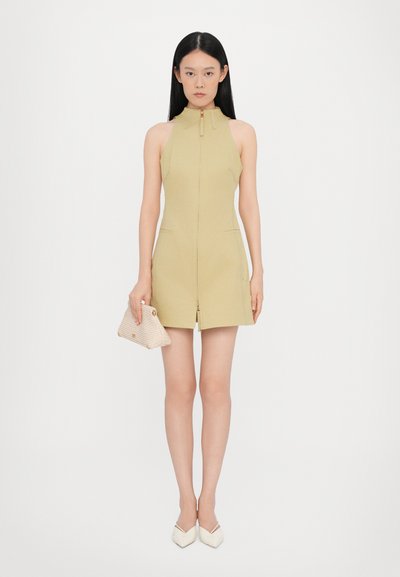 Knelang, ærmeløs kjole i lys gul med høj krave, lynlås foran og sidelommer. Modellen holder en beige clutch og bærer hvide hæle.
