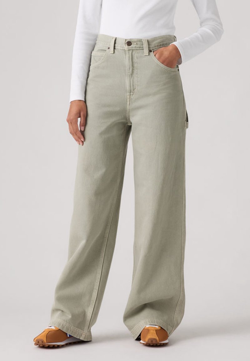 Personne portant un pantalon large beige clair, un haut à manches longues blanc et des baskets décontractées marron et blanches, debout avec une main dans la poche.
