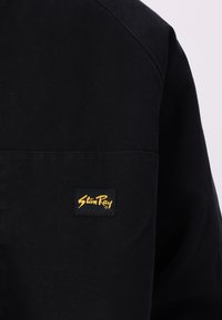Black fabric shirt featuring a rounded collar and a left chest pocket. A yellow "Stan Ray" label is stitched on the pocket.