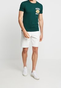 Camiseta verde oscura con el texto "RUGBY" y gráfico, combinada con pantalones cortos blancos y zapatillas blancas. Mangas cortas y corte clásico.