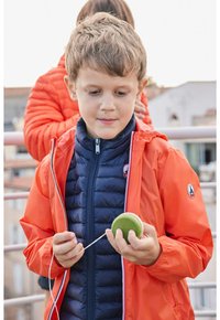 Chaqueta ligera de color naranja brillante superpuesta sobre una chaqueta acolchada azul marino. El niño sostiene un objeto verde con un cordón blanco, concentrado en el objeto.
