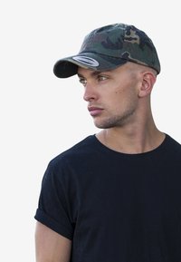 Căciulă de baseball cu camuflaj, cu o margine curbată și o etichetă albă. Purtați cu un tricou negru cu gât rotund, cu mâneci scurte suflate.