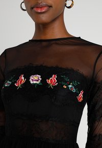 Robe longue transparente noire à manches longues avec broderie florale rouge, rose et verte, et accents en dentelle le long du col et des manches.