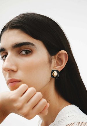 Pendientes cuadrados en oro y negro con un acento redondo central, con un acabado texturizado, exhibidos sobre un fondo neutro.