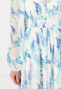 Robe blanche avec motifs floraux bleus et verts, manches longues transparentes, taille froncée et décolleté en V, montrée de la taille jusqu'à mi-cuisse.