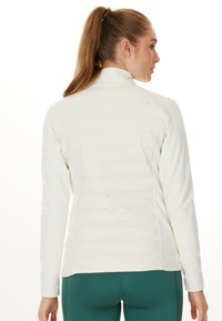 Lättviktig, krämfärgad jacka med kviltad rygg, figursydd design och långa ärmar, kombinerad med tealfärgade leggings.
