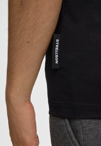 Zwarte T-shirt met een slim fit, geribbelde textuur en een zijkraag met "STRELLSON" in het wit. Grijze, textuurlijke broek gedeeltelijk zichtbaar.