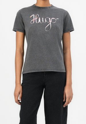 Osoba ubrana w wyblakłą, czarną koszulkę z krótkim rękawem z różowym napisem „Hugo” na piersi oraz czarne spodnie.