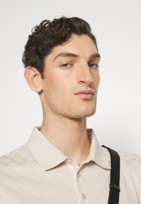 Polo beige con un patrón floral texturizado y un cuello clásico. El modelo lleva una bandolera negra. Fondo neutro.