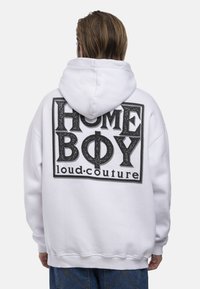 Balta kapuce ar lielu melnu grafikas druku aizmugurē, kurā ir teksts "HOME BOY loud.couture." Mīksts, biezs audums, atbrīvota piegriezuma.