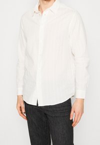 Chemise blanche à boutons avec des rayures verticales, des manches longues et un col classique, présentant une coupe décontractée et un tissu texturé léger.