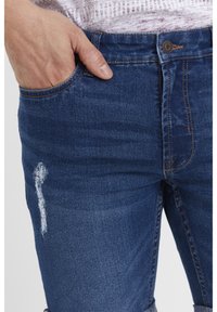 Mörkblå denimjeans med femficksdesign, synliga sömmar och en sliten detalj på låret. Knäppning med knapp i midjan.