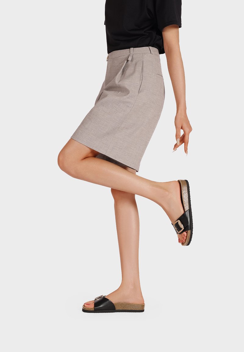 Gonna grigia su misura con pieghe, abbinata a sandali neri con fibbia e suola testurizzata. Il modello posa con una gamba sollevata.