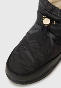 Chinelo preto acolchoado com bico arredondado, apresentando um botão decorativo dourado e um fecho de cordão na parte superior. Forro interior macio visível.
