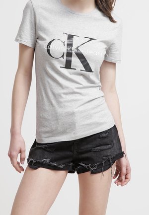 Persoon draagt een lichtgrijs Calvin Klein Jeans T-shirt en zwarte versleten denim shorts tegen een effen achtergrond.