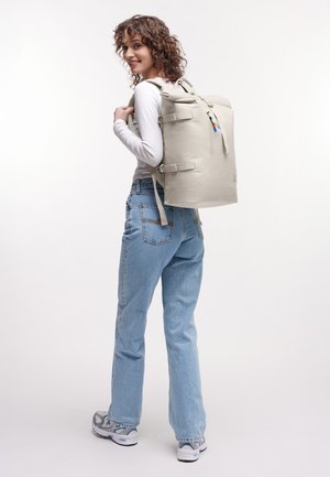 Donna con capelli ricci che indossa una camicia bianca a maniche lunghe, jeans azzurri chiari e scarpe da ginnastica, con uno zaino beige grande sulle spalle.