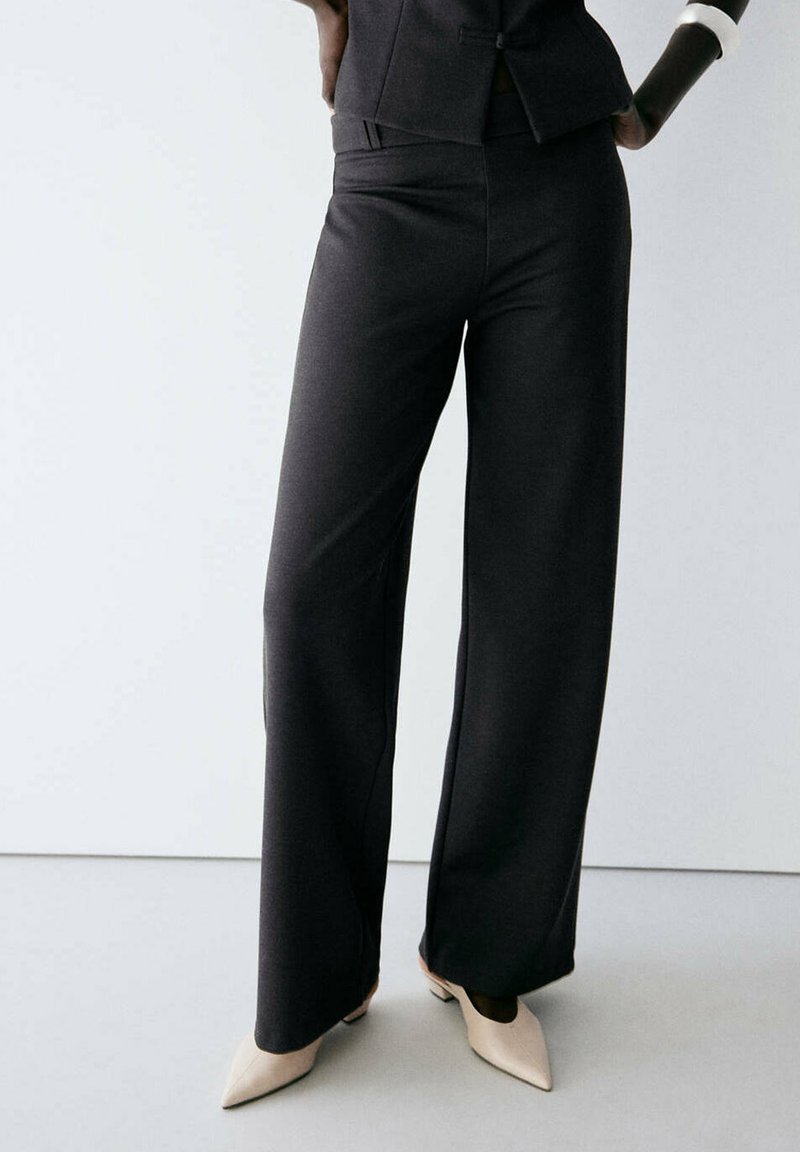 Personne portant un pantalon noir taille haute à jambes larges et des talons blancs à bouts pointus, debout devant un fond clair uni.