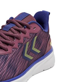 Fioletowy but sportowy z teksturowaną siateczkową cholewką i gładkimi nakładkami. Posiada niebieskie akcenty i żółte paski w kształcie jodełki. Biała podeszwa zewnętrzna.