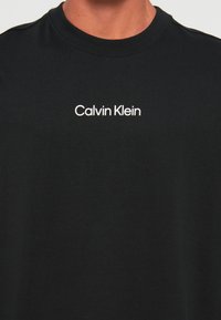 Sort crew neck t-shirt med hvid "Calvin Klein" logo centreret på brystet, båret af en person med synlig hals og overkrop.