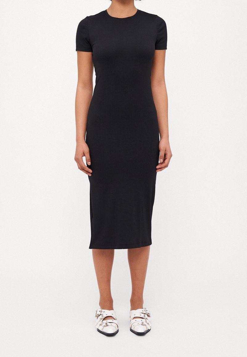 Robe midi noire à côtes, à manches courtes, avec col rond et fentes sur les côtés. Portée avec des chaussures blanches à bout ouvert et à boucle.