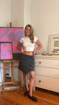 Weißes, kurzes T-Shirt mit gestickten Details, grauer Wickelrock mit Knopf und schwarze Slipper auf einem Holzboden, im Hintergrund abstrakte Kunst und Möbel.