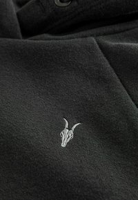 Felpa nera con una texture morbida, caratterizzata da un piccolo design ricamato di teschio di capra in grigio chiaro. Include un dettaglio con colletto a bottoni.