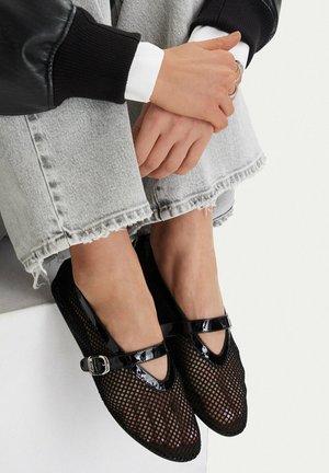 Feet wearing black mesh Mary Jane flats with a buckle, paired with frayed hem light gray jeans and hands resting on knees.