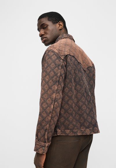 Άνδρας που φοράει καφέ τζιν μπουφάν με σχέδιο και λεπτομέρεια στο γιόκ, κοιτάζοντας πάνω από τον αριστερό του ώμο σε απλό φωτεινό φόντο.