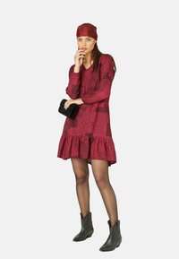 Rotes Kleid mit ausgestellt geschnittenem Saum, langen Ärmeln und schwarzem Punktmuster; kombiniert mit einem passenden Kopftuch und einer schwarzen Clutch. Graue knöchelhöhe Stiefel.