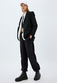 Schwarzes oversized Blazer, weißes Hemd mit Knöpfen, schwarzes Cropped-Top, lockere schwarze Hose und schwarze Kampfstiefel. Weißer Hut als Accessoire.