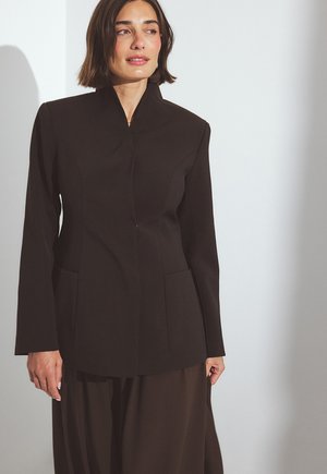 Musta räätälöity takki, jossa on korkea kaulus, pitkät hihat ja kaksi etutaskua. Silkkinen kangas, jossa on jämäkkä istuvuus ja minimalistinen muotoilu.