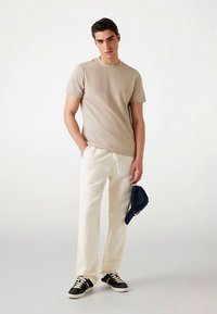 Beige geribd T-shirt, crèmekleurige broek en zwarte sneakers. De persoon houdt een blauwe pet met een wit logo vast. Simpele, casual outfit.