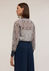 Průhledná vzorová blůza s geometrickým designem ve světle modré a béžové barvě, s dlouhými rukávy a kontrastními pruhovanými manžetami. V kombinaci s námořnickými kalhotami.