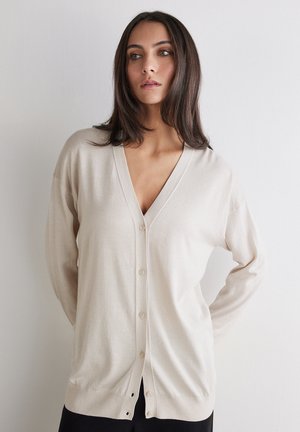Donna con capelli scuri che indossa un cardigan beige abbottonato, in piedi con le mani dietro la schiena, guardando leggermente di lato contro una parete neutra.