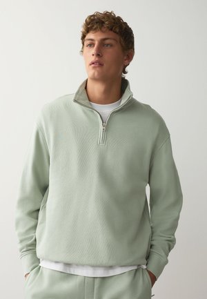 Junger Mann mit lockigem Haar trägt ein hellgrünes Sweatshirt mit Viertelreißverschluss und passende Hose, steht mit den Händen in den Taschen vor einem schlichten Hintergrund.