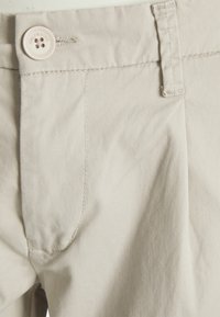 Light beige cotton pants with a smooth texture, featuring a front pocket and a large button with branding. Tailored design with precise stitching.